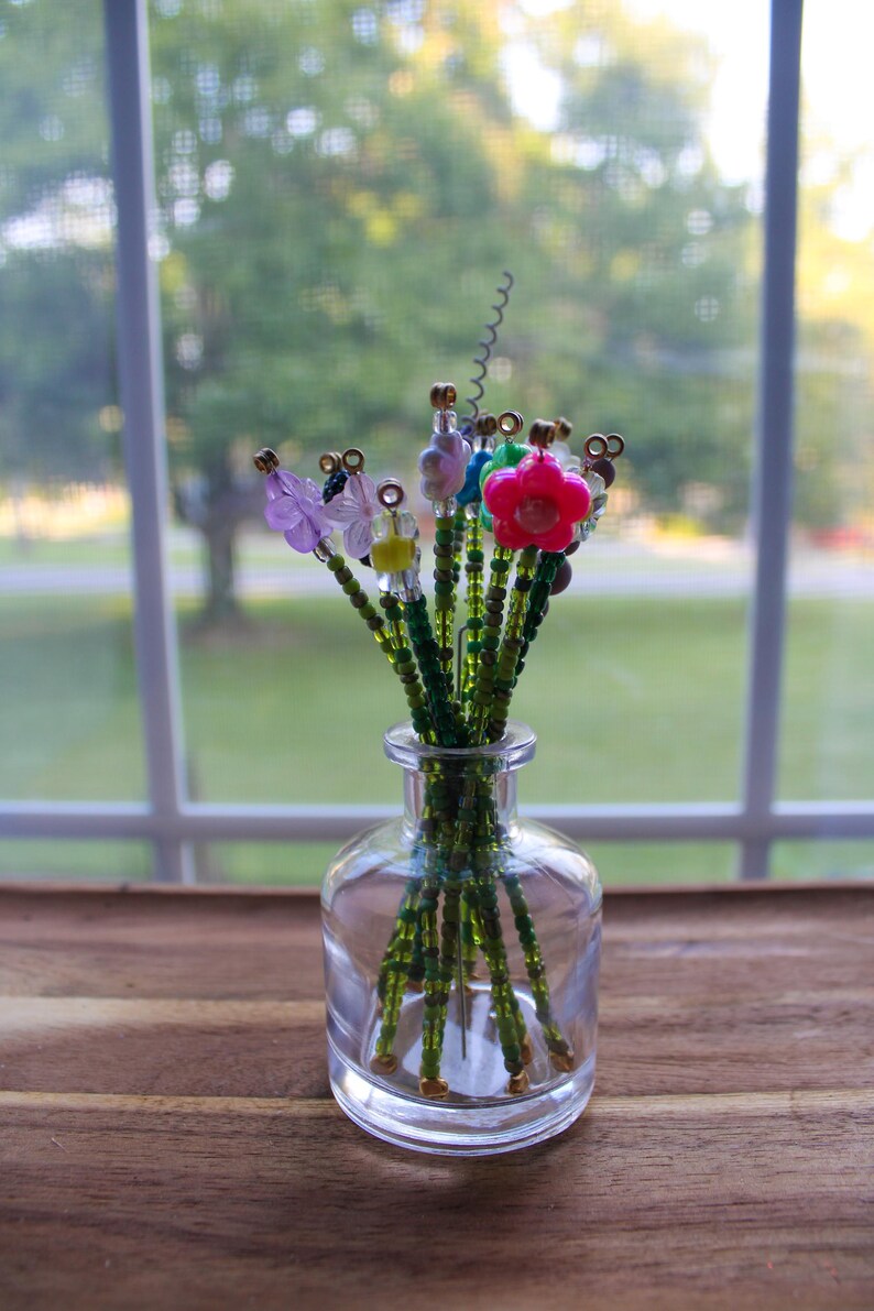 May include: A clear glass vase holding a bouquet of beaded flowers. The flowers have green stems and colorful beaded petals in pink, purple, blue, and yellow. The vase sits on a wooden surface.