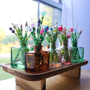 May include: A wooden tray holds several small glass vases, each containing beaded floral arrangements. The vases are various colors, including green, amber, and clear. The beaded flowers are colorful, with pink, blue, and purple accents.