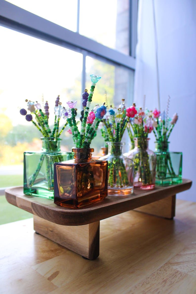 May include: A wooden tray holds several small glass vases, each containing beaded floral arrangements. The vases are various colors, including green, amber, and clear. The beaded flowers are colorful, with pink, blue, and purple accents.