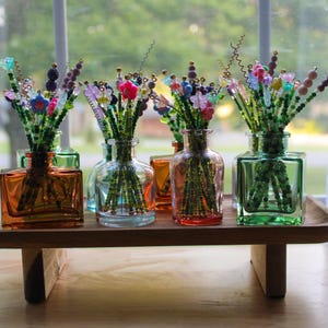 May include: A wooden tray holds several glass vases filled with beaded floral arrangements. The vases are square or rounded, in shades of orange, green, and blue. The beaded flowers have colorful accents.
