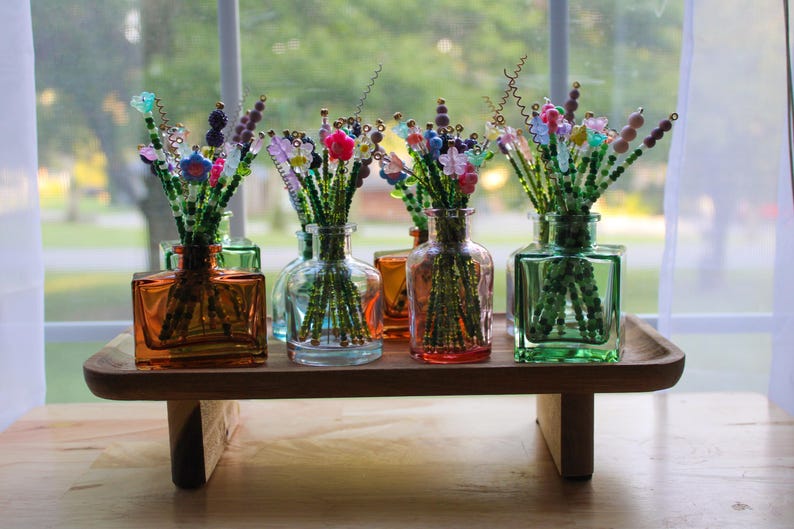 May include: A wooden tray holds several glass vases filled with beaded floral arrangements. The vases are square or rounded, in shades of orange, green, and blue. The beaded flowers have colorful accents.