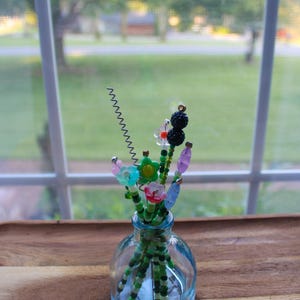 May include: A small, clear glass vase holding a collection of beaded flowers. The flowers are made of colorful beads in various shapes, including flowers and berries. The vase sits on a wooden surface, with a window and greenery in the background.