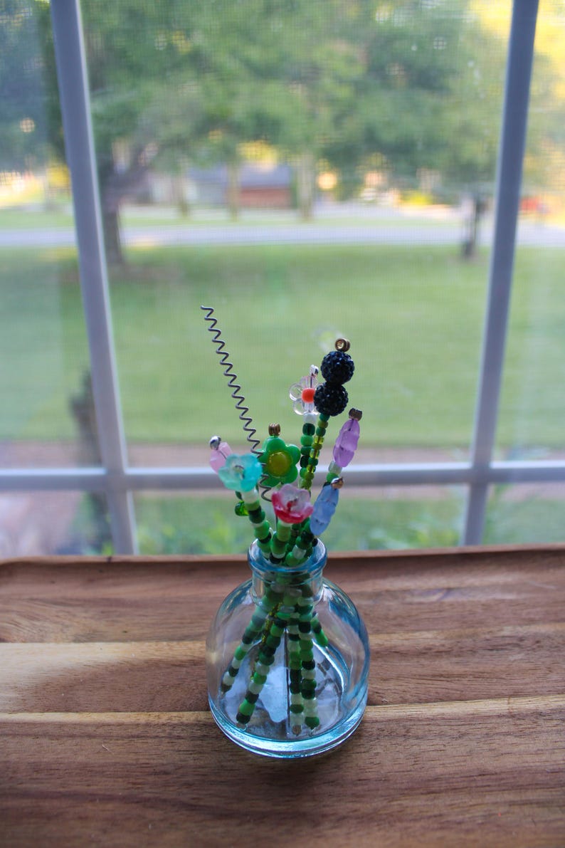 May include: A small, clear glass vase holding a collection of beaded flowers. The flowers are made of colorful beads in various shapes, including flowers and berries. The vase sits on a wooden surface, with a window and greenery in the background.