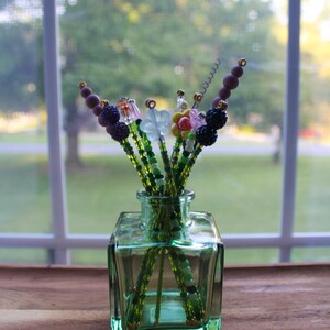 May include: A small, square, green glass vase holding beaded floral stems. The stems are green with various colorful beaded flower and berry accents. The vase sits on a wooden surface in front of a window.