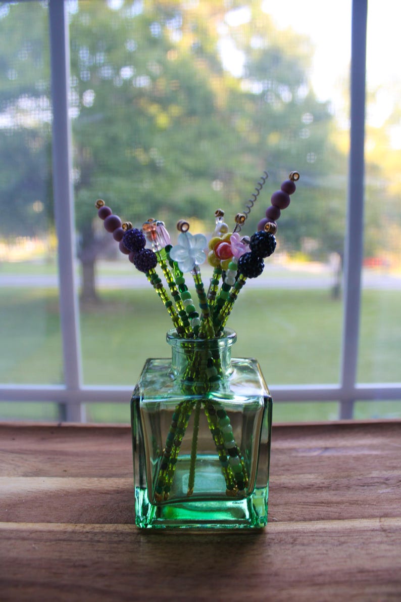 May include: A small, square, green glass vase holding beaded floral stems. The stems are green with various colorful beaded flower and berry accents. The vase sits on a wooden surface in front of a window.