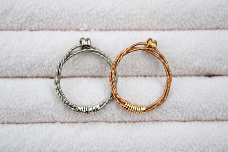May include: Two guitar string bracelets, one silver and one gold, on a white background.