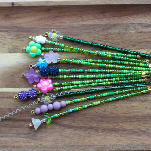 May include: A collection of beaded hair pins with floral and spherical accents. The pins feature a variety of colors, including green, pink, purple, and blue, with gold-colored metal accents. The beads are arranged in a linear pattern.