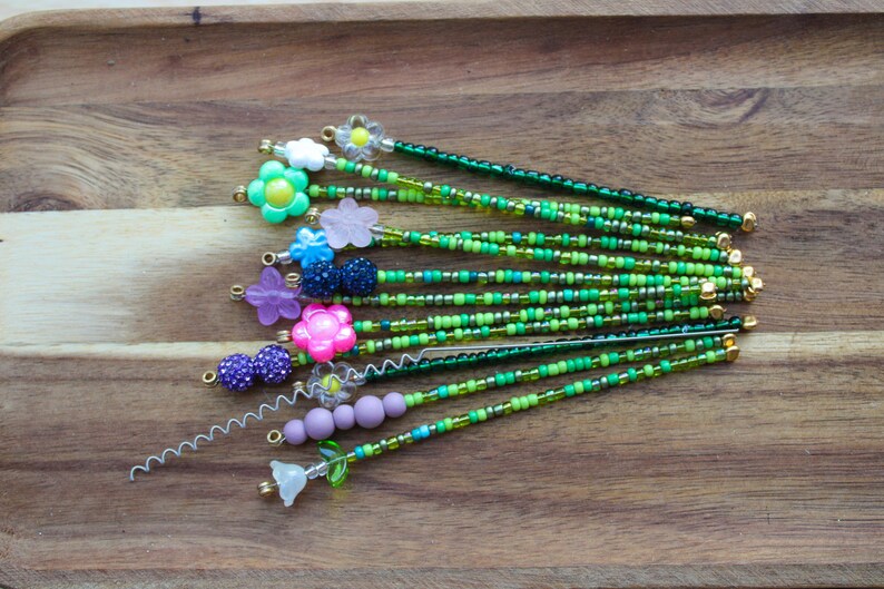 May include: A collection of beaded hair pins with floral and spherical accents. The pins feature a variety of colors, including green, pink, purple, and blue, with gold-colored metal accents. The beads are arranged in a linear pattern.
