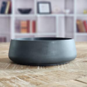 May include: A dark gray, low-profile bowl with a rounded shape. The bowl is made of a matte material and sits on a light brown wooden surface. The background is blurred, showing a white shelf with books and decorative items.