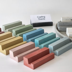 May include: A set of ten concrete display stands in various colors, including red, pink, yellow, green, blue, and gray. The stands are arranged in a row on a white surface. The stands are all the same size and shape, and they have a small lip on the front to hold items in place. The stands are labeled "Casting Theory".