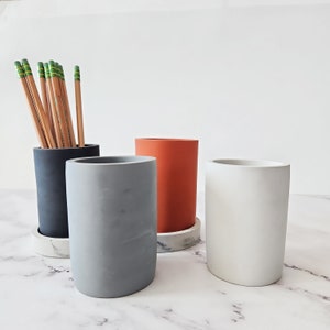 May include: Three concrete pencil holders in gray, white, and orange with a marble base. The gray holder is in the foreground, the white holder is in the background, and the orange holder is in the middle. The gray holder has a pencil holder with a bunch of pencils in it.