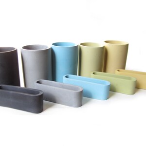 Op de afbeelding: Een set van acht betonnen containers in verschillende kleuren, waaronder grijs, blauw, groen en geel. De containers zijn in een verspringende opstelling geplaatst, met de grootste container aan de achterkant en de kleinste container aan de voorkant. Elke container heeft een rechthoekige vorm met een afgeronde rand.