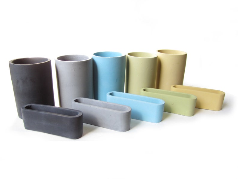 Op de afbeelding: Een set van acht betonnen containers in verschillende kleuren, waaronder grijs, blauw, groen en geel. De containers zijn in een verspringende opstelling geplaatst, met de grootste container aan de achterkant en de kleinste container aan de voorkant. Elke container heeft een rechthoekige vorm met een afgeronde rand.