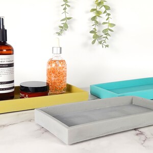 May include: Three rectangular concrete trays in yellow, gray, and turquoise blue. The trays are arranged in a staggered formation on a white marble surface. A brown glass bottle with a black spray top and a clear glass bottle with a dropper top are on the yellow tray. A small round jar with a black lid is also on the yellow tray.