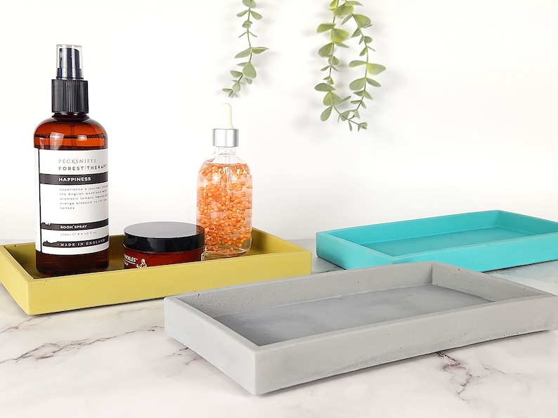 May include: Three rectangular concrete trays in yellow, gray, and turquoise blue. The trays are arranged in a staggered formation on a white marble surface. A brown glass bottle with a black spray top and a clear glass bottle with a dropper top are on the yellow tray. A small round jar with a black lid is also on the yellow tray.