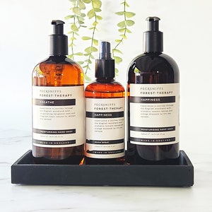 May include: Three amber glass bottles with black pump tops and labels. The bottles are arranged in a row on a black tray. The labels read "Pecksniffs Forest Therapy" and feature the scents "Breathe", "Happiness", and "Happiness" (room spray).
