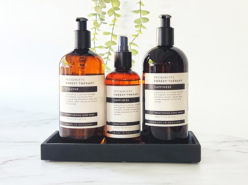 May include: Three amber glass bottles with black pump tops and labels. The bottles are arranged in a row on a black tray. The labels read "Pecksniffs Forest Therapy" and feature the scents "Breathe", "Happiness", and "Happiness" (room spray).