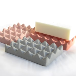 May include: Three soap dishes made of concrete with a textured, triangular pattern. The dishes are in shades of pink and gray. A white bar of soap is resting on top of the two pink dishes.