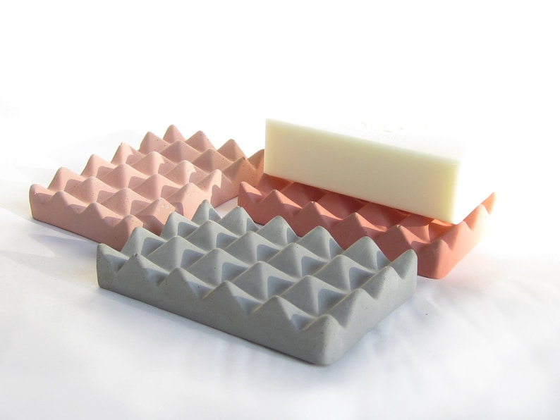 May include: Three soap dishes made of concrete with a textured, triangular pattern. The dishes are in shades of pink and gray. A white bar of soap is resting on top of the two pink dishes.
