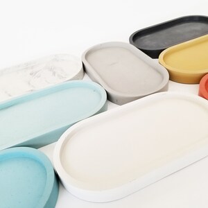 May include: A set of nine oval-shaped concrete trays in various colours, including white, light blue, grey, yellow, red, and black. The trays are arranged in a row, with the largest tray at the back and the smallest tray at the front.