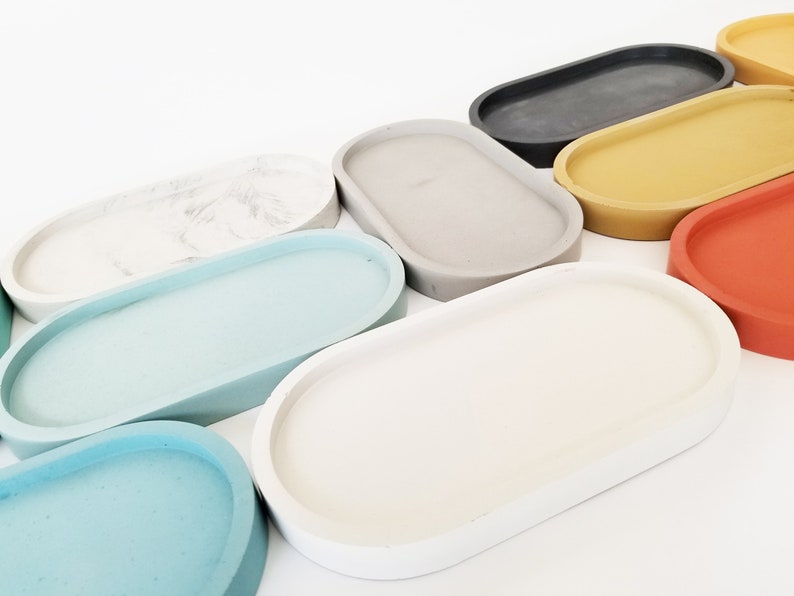 May include: A set of nine oval-shaped concrete trays in various colours, including white, light blue, grey, yellow, red, and black. The trays are arranged in a row, with the largest tray at the back and the smallest tray at the front.