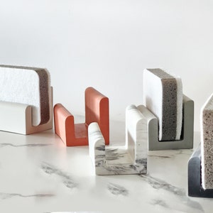 May include: Five different colored sponge holders with sponges in them. The holders are made of ceramic and are in the shapes of a U. The sponges are in various colors and textures.