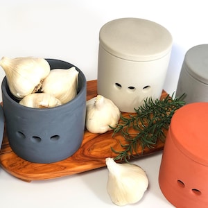 May include: Three ceramic garlic keepers with lids. The keepers are in grey, beige, and orange. The grey keeper is filled with garlic cloves. The keepers are on a wooden chopping board with a sprig of rosemary.