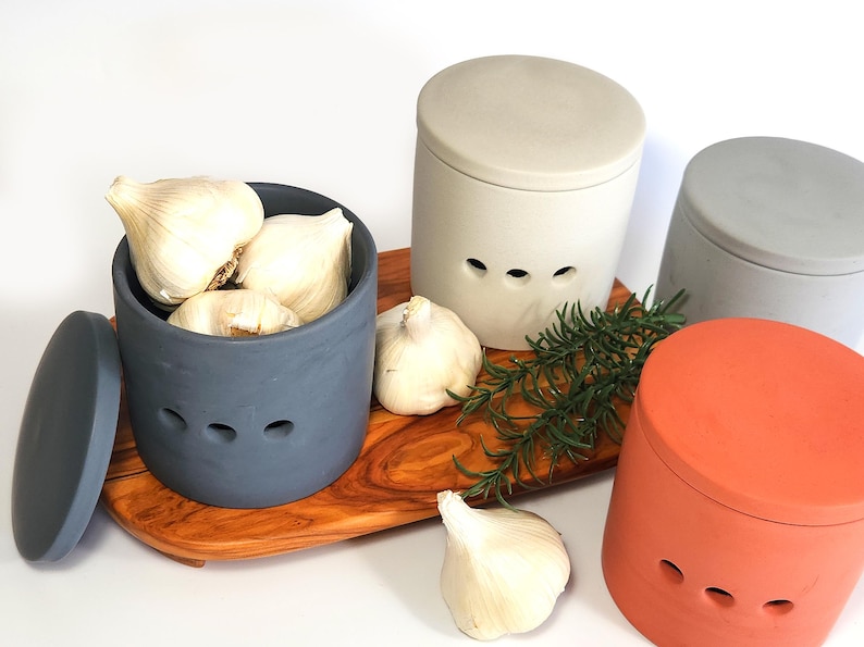 May include: Three ceramic garlic keepers with lids. The keepers are in grey, beige, and orange. The grey keeper is filled with garlic cloves. The keepers are on a wooden chopping board with a sprig of rosemary.