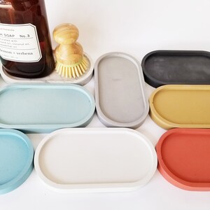 May include: A set of nine oval-shaped concrete trays in various colours, including teal, grey, black, yellow, and red. The trays are arranged in a semi-circle on a white surface.