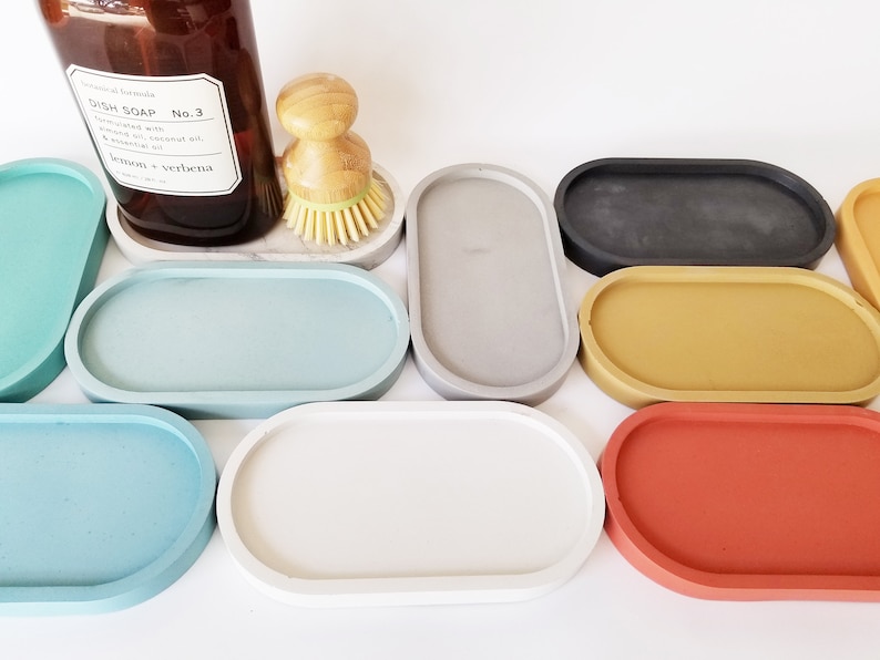 May include: A set of nine oval-shaped concrete trays in various colours, including teal, grey, black, yellow, and red. The trays are arranged in a semi-circle on a white surface.