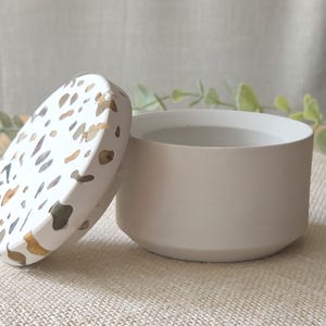 May include: A small, off-white ceramic container with a matching lid. The lid features a terrazzo design with brown, gray, and gold flecks. The container is cylindrical and sits on a neutral-colored surface.