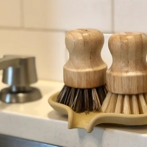 May include: Two wooden-handled dish brushes with natural bristles, sitting in a beige ceramic dish soap holder.