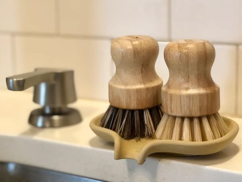 May include: Two wooden-handled dish brushes with natural bristles, sitting in a beige ceramic dish soap holder.