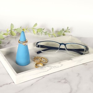 May include: A white marble tray with a blue ring holder and gold rings. A pair of blue-rimmed glasses is resting on the tray.