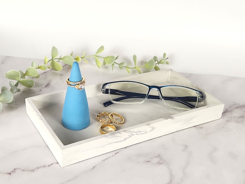May include: A white marble tray with a blue ring holder and gold rings. A pair of blue-rimmed glasses is resting on the tray.