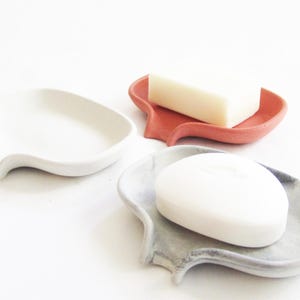 May include: Three ceramic soap dishes in different colors: white, coral, and gray. Each dish has a unique, organic shape with a curved lip and a small spout. The dishes are designed to hold bars of soap.