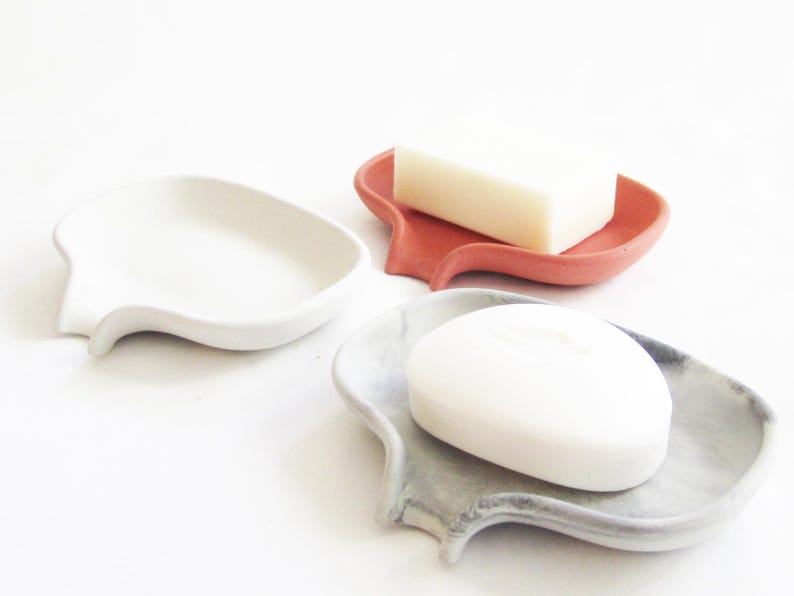 May include: Three ceramic soap dishes in different colors: white, coral, and gray. Each dish has a unique, organic shape with a curved lip and a small spout. The dishes are designed to hold bars of soap.