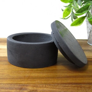 May include: A black concrete ring dish with a lid. The dish is round and has a smooth surface. The lid is also round and has a smooth surface.