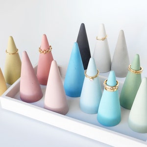 Pu&ograve; includere: Una collezione di dodici porta anelli a forma di cono in vari colori pastello e neutri, tra cui giallo, rosa, blu, verde, grigio e nero. Alcuni coni sono sormontati da anelli in metallo dorato, il tutto disposto su un vassoio rettangolare bianco.
