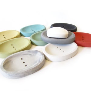 Puede incluir: Surtido de jaboneras ovaladas en varios colores, incluyendo verde, azul, gris, blanco y rojo. Las jaboneras est&aacute;n hechas de hormig&oacute;n y tienen tres agujeros de drenaje. Una pastilla de jab&oacute;n blanca descansa sobre una de las jaboneras.