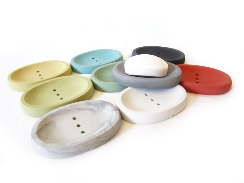 Puede incluir: Surtido de jaboneras ovaladas en varios colores, incluyendo verde, azul, gris, blanco y rojo. Las jaboneras est&aacute;n hechas de hormig&oacute;n y tienen tres agujeros de drenaje. Una pastilla de jab&oacute;n blanca descansa sobre una de las jaboneras.
