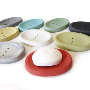 Puede incluir: Surtido de jaboneras ovaladas en varios colores, incluyendo gris, amarillo, verde, azul, blanco, negro y rojo. Las jaboneras est&aacute;n hechas de hormig&oacute;n y tienen tres agujeros de drenaje. Una pastilla de jab&oacute;n blanca est&aacute; sobre una jabonera roja.