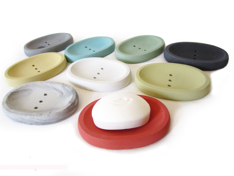 Puede incluir: Surtido de jaboneras ovaladas en varios colores, incluyendo gris, amarillo, verde, azul, blanco, negro y rojo. Las jaboneras est&aacute;n hechas de hormig&oacute;n y tienen tres agujeros de drenaje. Una pastilla de jab&oacute;n blanca est&aacute; sobre una jabonera roja.