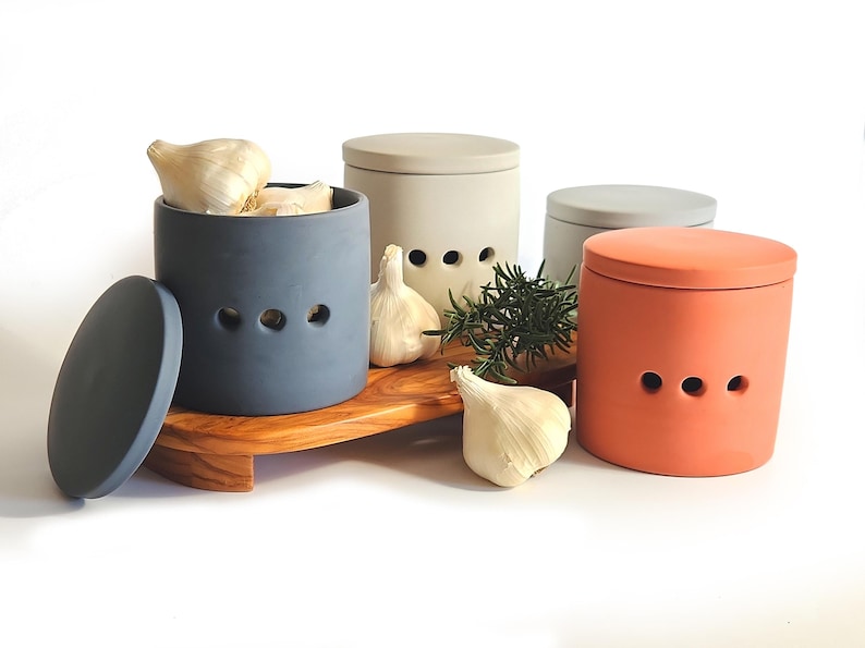 May include: Four ceramic garlic keepers with lids, three in shades of grey and one in a terracotta orange. The keepers have holes for air circulation and are arranged on a wooden chopping board with fresh garlic cloves and rosemary sprigs.