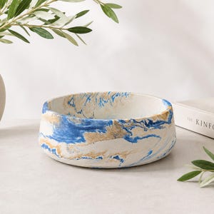 May include: A round, shallow bowl featuring a marbled pattern of white, vibrant blue, and metallic gold swirls. The bowl has a smooth, matte finish and a wide, open top, showcasing a continuous, fluid design that wraps around the exterior surface.