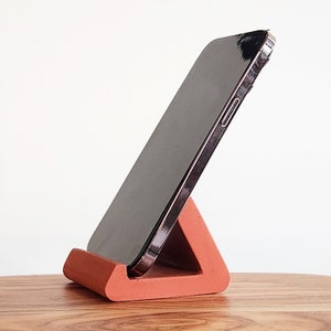 May include: A terracotta phone stand in the shape of a triangle, holding a black smartphone in a vertical position.