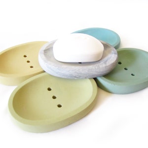 Puede incluir: Conjunto de cinco jaboneras ovaladas en varios tonos de verde y amarillo. Las jaboneras est&aacute;n hechas de hormig&oacute;n y tienen agujeros de drenaje. Una pastilla de jab&oacute;n blanca descansa sobre una de las jaboneras.