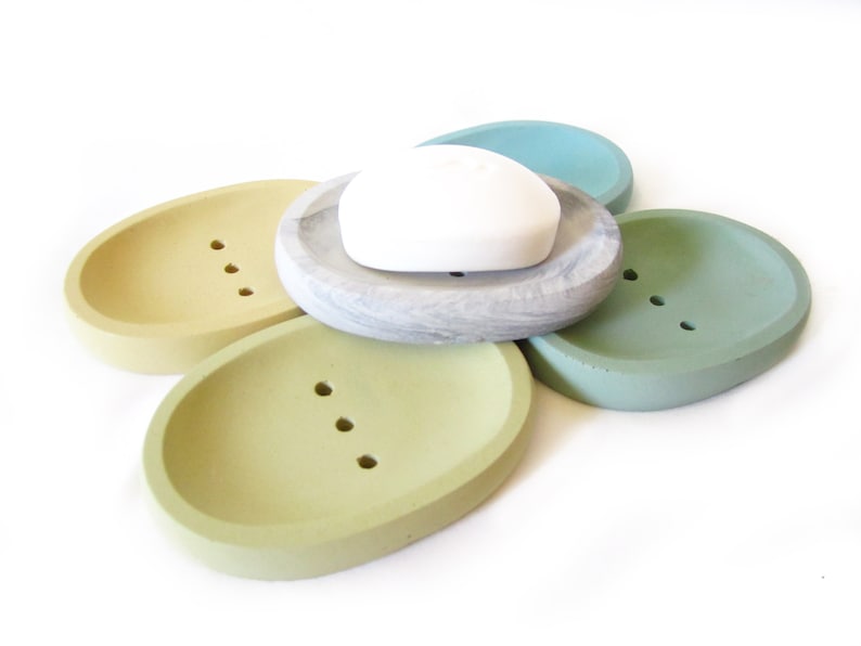 Puede incluir: Conjunto de cinco jaboneras ovaladas en varios tonos de verde y amarillo. Las jaboneras est&aacute;n hechas de hormig&oacute;n y tienen agujeros de drenaje. Una pastilla de jab&oacute;n blanca descansa sobre una de las jaboneras.