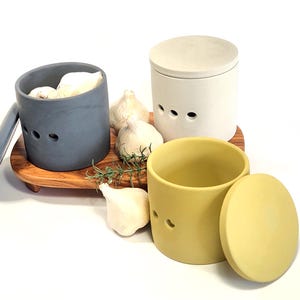 May include: Three ceramic garlic keepers with lids. One is grey, one is white, and one is yellow. The keepers are on a wooden tray with garlic cloves and rosemary sprigs.