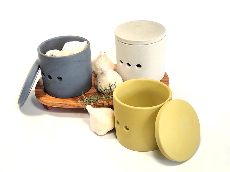 May include: Three ceramic garlic keepers with lids. One is grey, one is white, and one is yellow. The keepers are on a wooden tray with garlic cloves and rosemary sprigs.